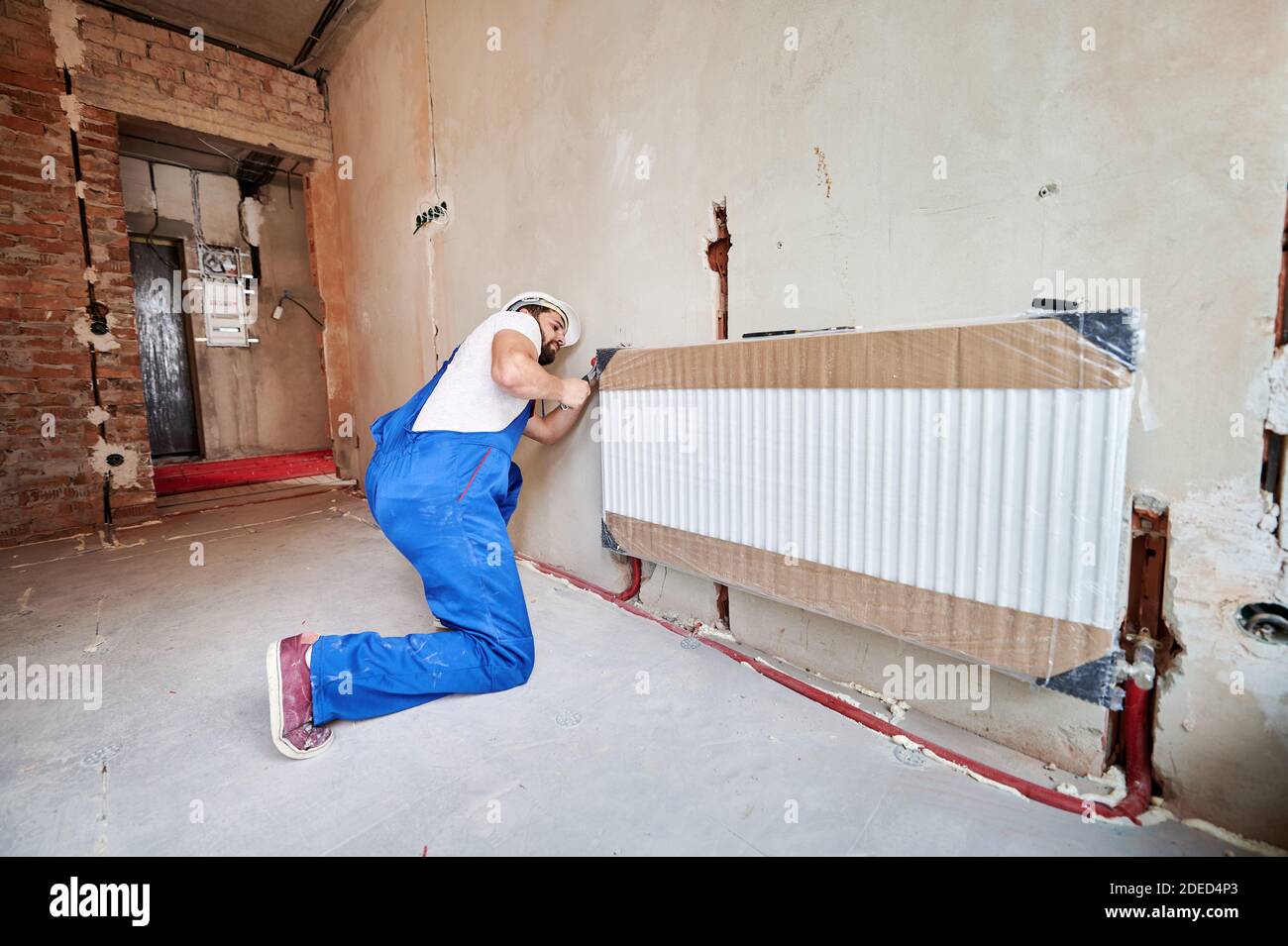 Plumber in work overalls using wrench while installing heating radiator in room. Man in helmet