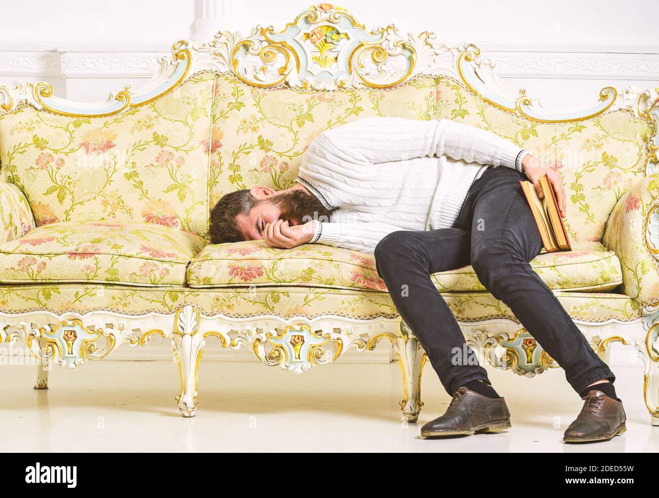 Macho sleep with book in hand. Boring literature concept. Man with beard  and mustache lay on baroque style sofa, holds book, white wall background.  Guy fall asleep while reading old boring book