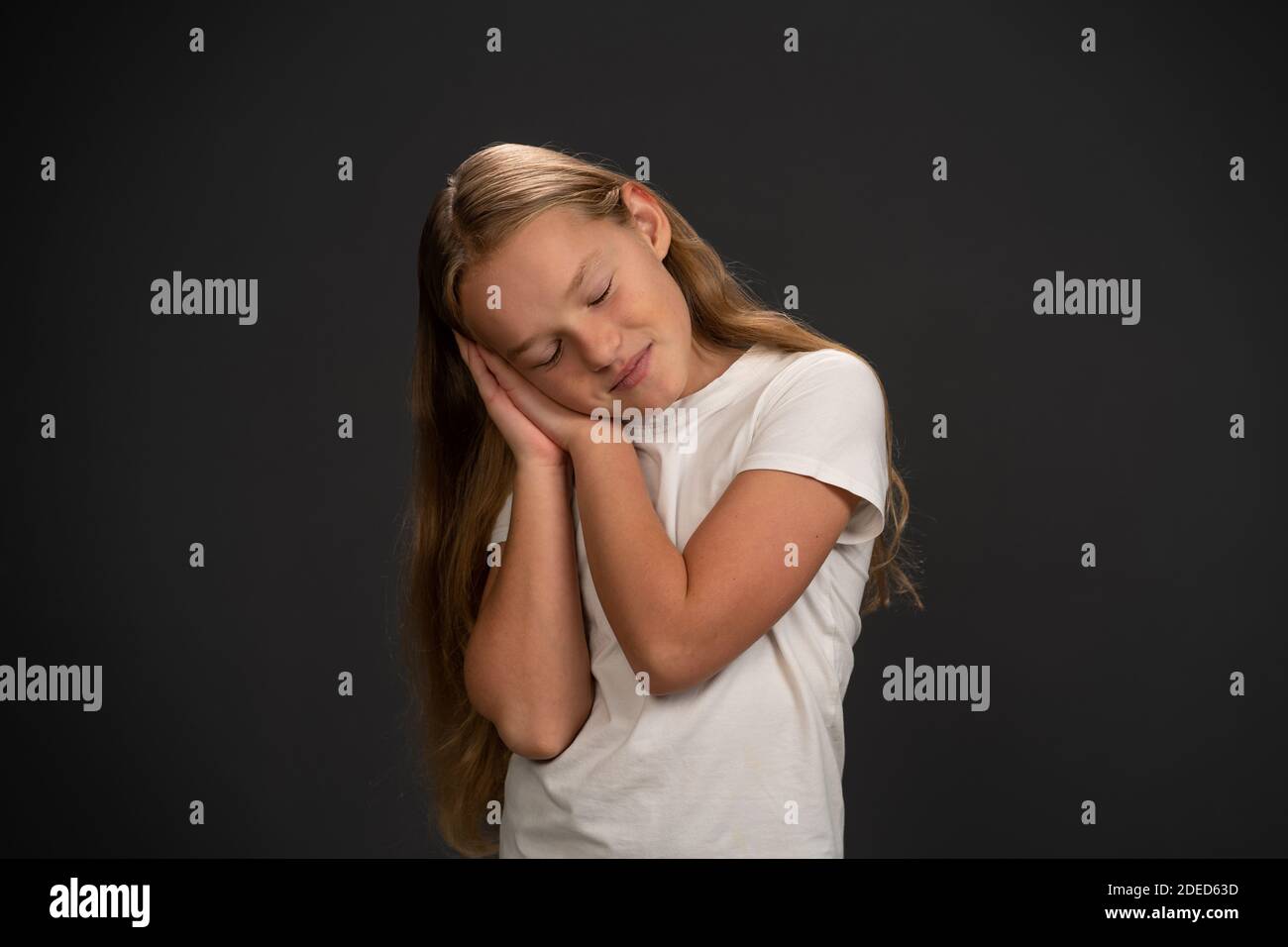 Going to sleep 8,10 years old girl holding her hands together and leaned head on them showing she is asleep wearing white t shirt smiling at the Stock Photo