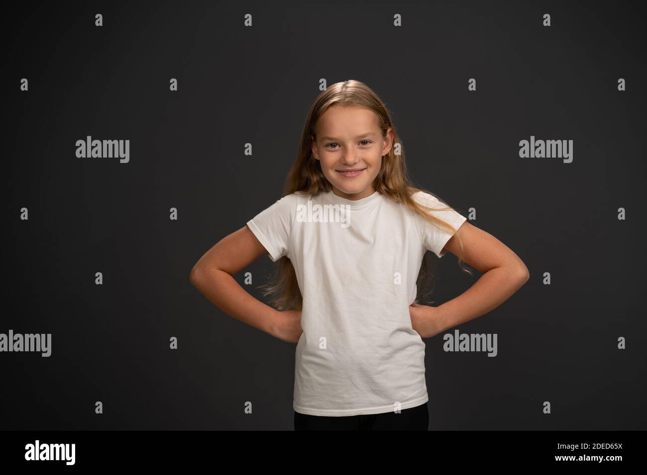Bossy little girl of 8,10 years put her hands sideways looks questioningly at the camera wearing white t shirt isolated on dark grey or black Stock Photo