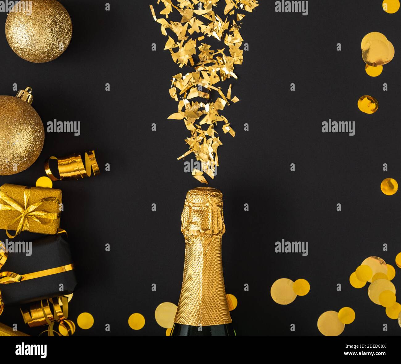 Golden bottle of champagne spills out gold sparkles in frame of gold festive Christmas decor confetti balls gifts on black background. Flat lay New Stock Photo