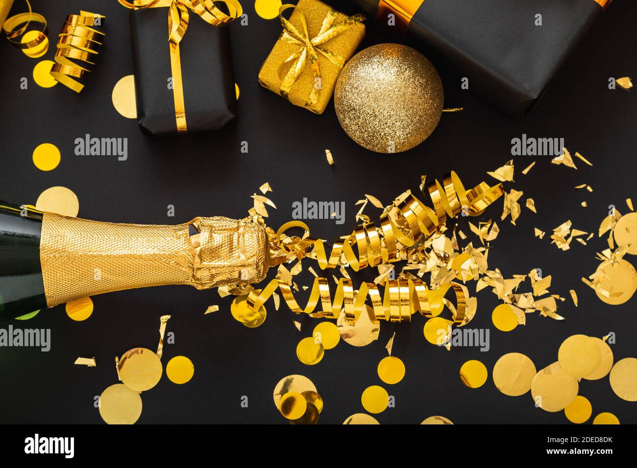 Golden bottle of champagne spills out gold sparkles in frame of gold festive Christmas decor confetti balls gifts on black background. Flat lay New Stock Photo