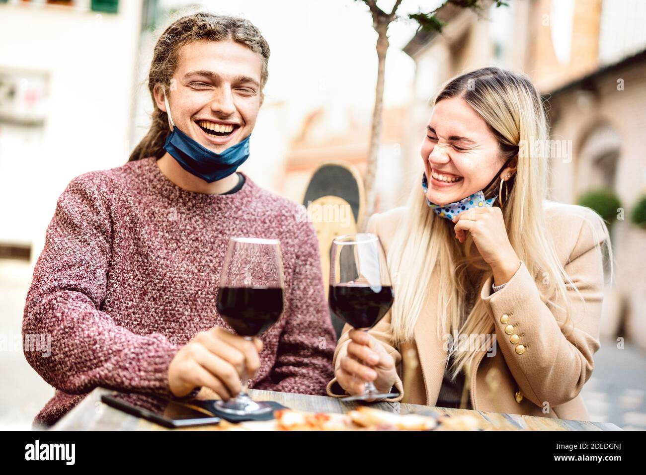 Young couple in love with open face masks having fun at wine bar outdoors - Happy millenial lovers enjoying lunch together at restaurant patio Stock Photo