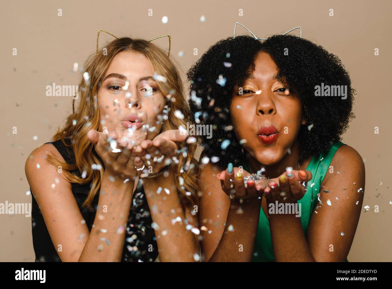 Black and Caucasian beautiful young women blowing confetti into the camera on a beige background. Celebrating Christmas in the studio. Party, event and celebration concept. Stock Photo