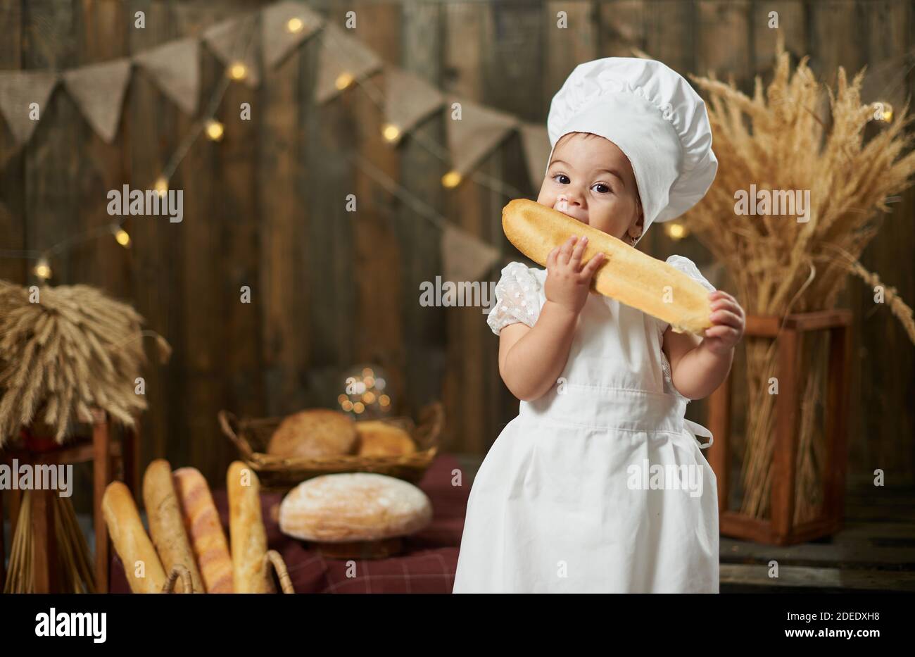 Cute baby girl wearing baker costume eating long bread Stock Photo