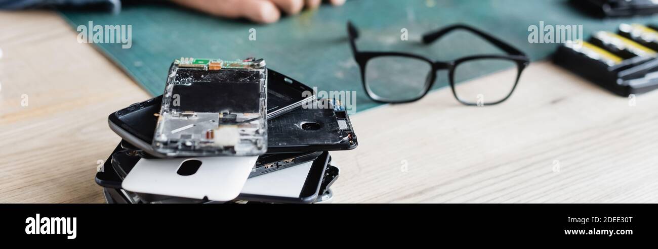 Close up view of pile of broken mobile phones near eyeglasses on workplace with blurred repairman on background, banner Stock Photo