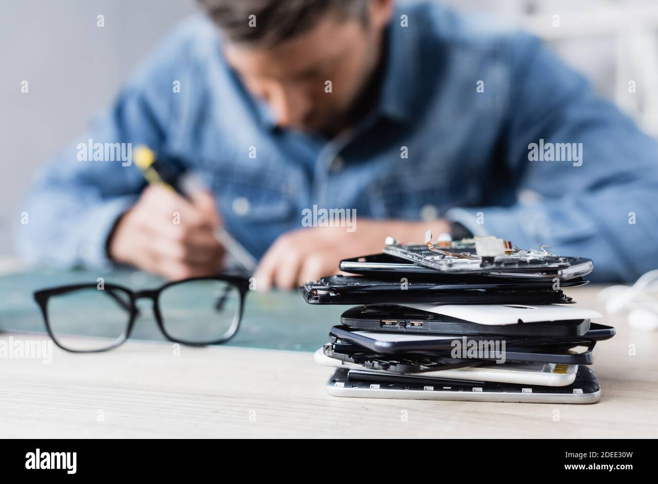 Close up view of pile of broken mobile phones near eyeglasses on workplace with blurred repairman on background Stock Photo