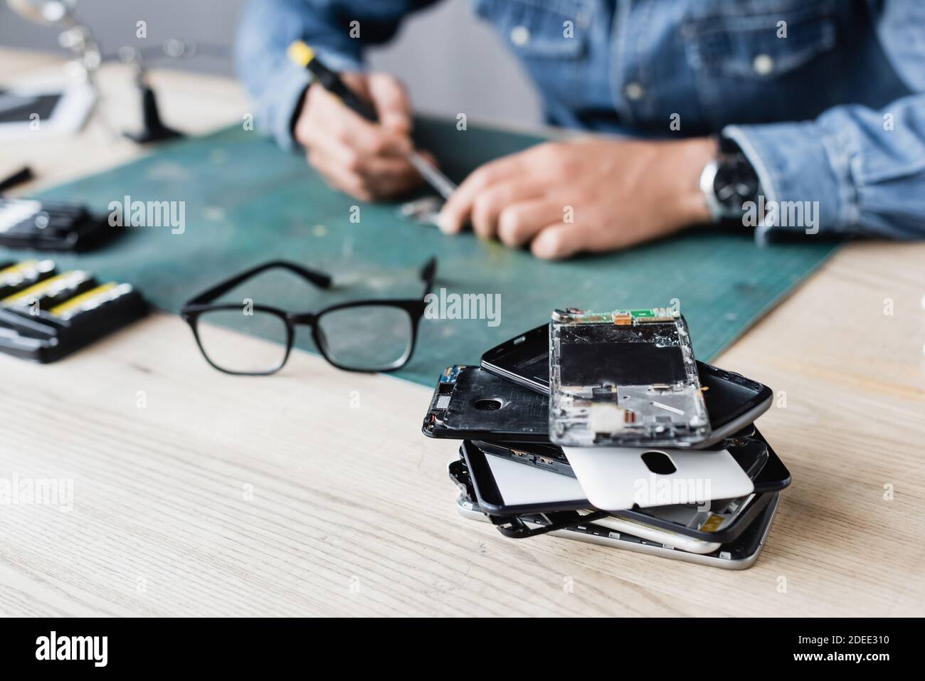 Close up view of pile with broken mobile phones near eyeglasses on workplace with blurred repairman working on background Stock Photo