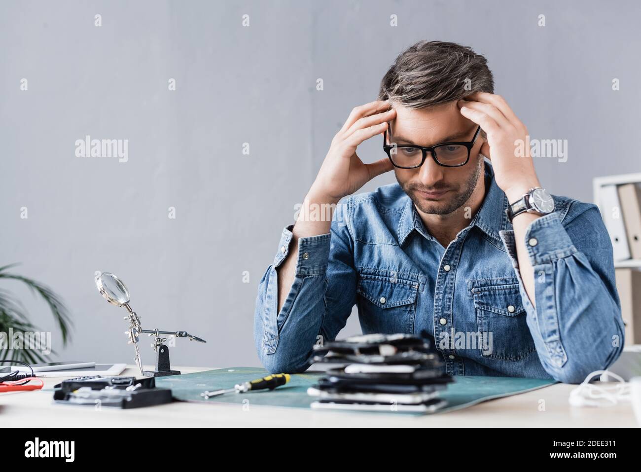 Tired repairman with hands near head looking at pile of broken mobile phones on blurred foreground Stock Photo
