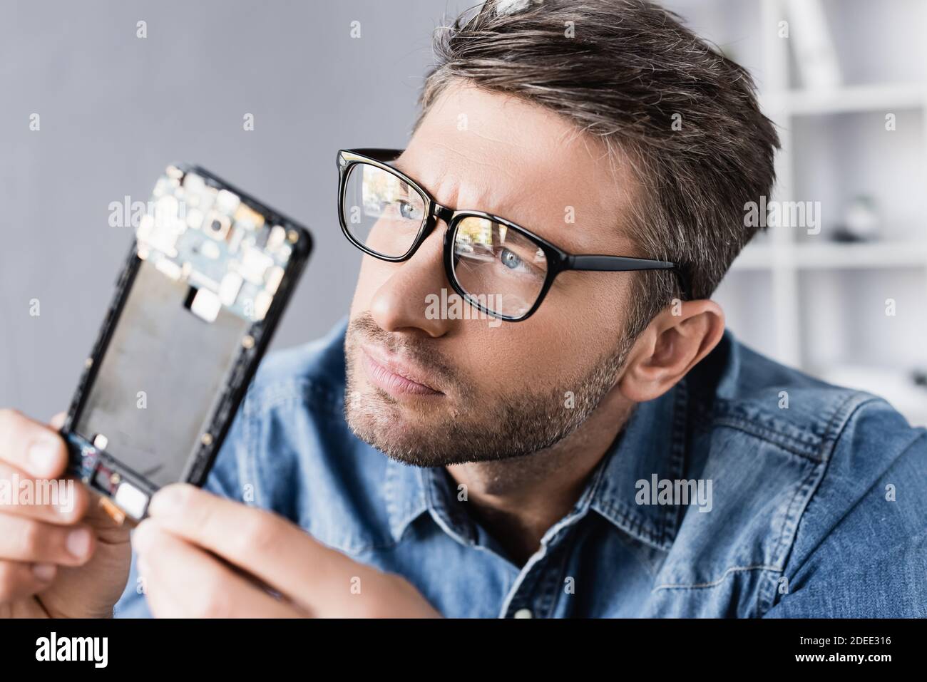 Concentrated repairman in eyeglasses looking at disassembled part of mobile phone on blurred foreground Stock Photo