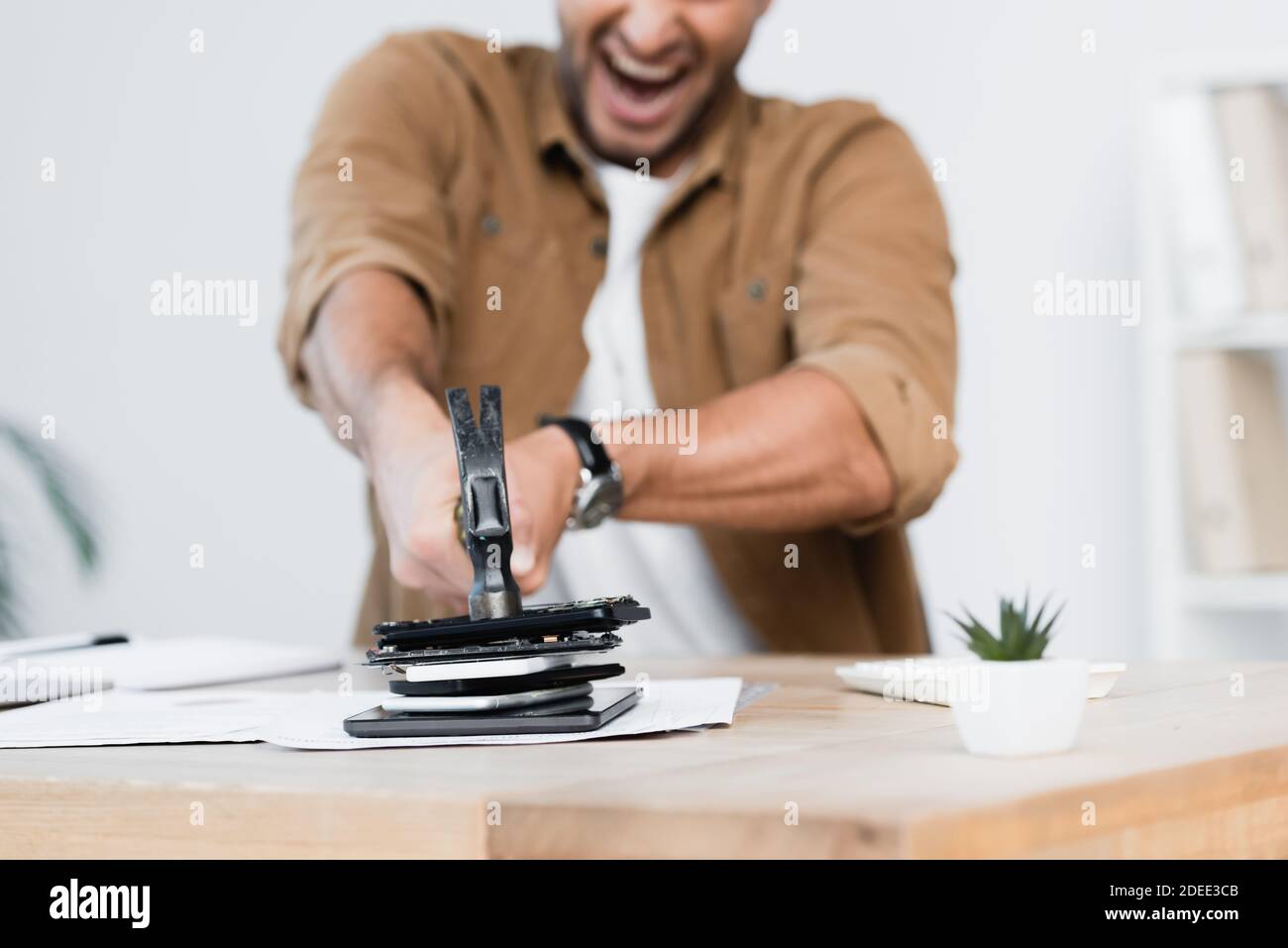Cropped view of shouting businessman cracking pile of smartphones with hammer on table on blurred background Stock Photo