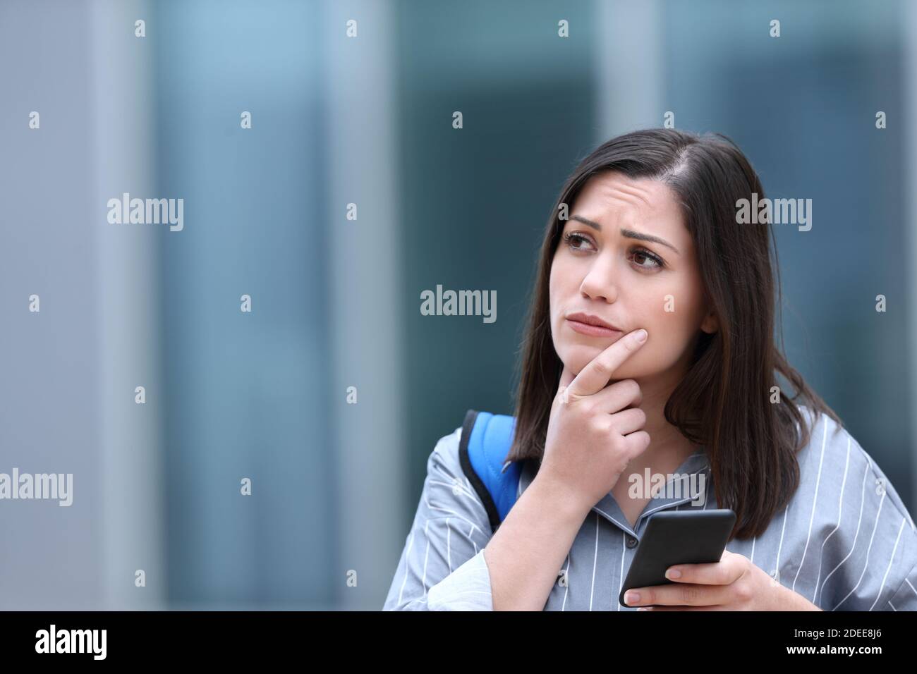 Worried student holding smart phone thinking looking at side in the street Stock Photo