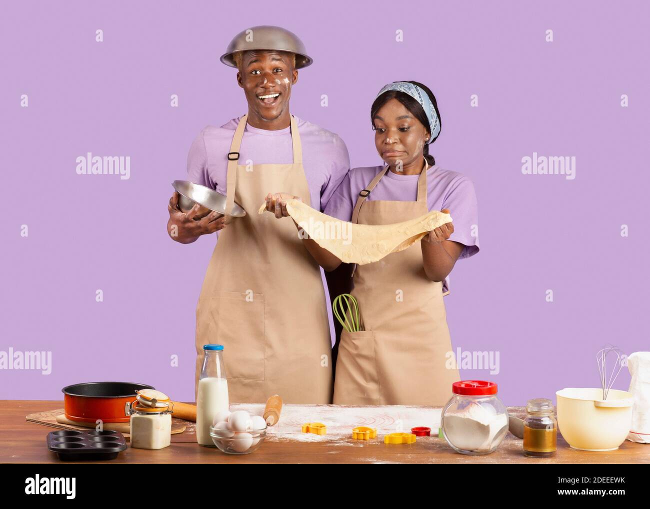 Positive black couple in aprons having dirty faces, holding cookie dough, baking together on lilac studio background Stock Photo