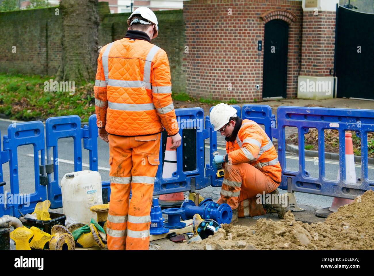 Two men wearing high visibility clothing carrying out road works for water and gas. One man kneeling one man supervising standing. Using power tools. Stock Photo