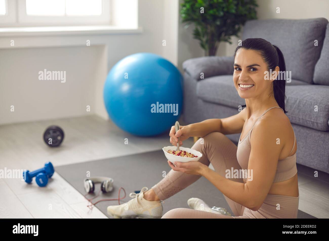 Woman sitting on sports mat and smiling at camera while enjoying healthy meal after workout Stock Photo