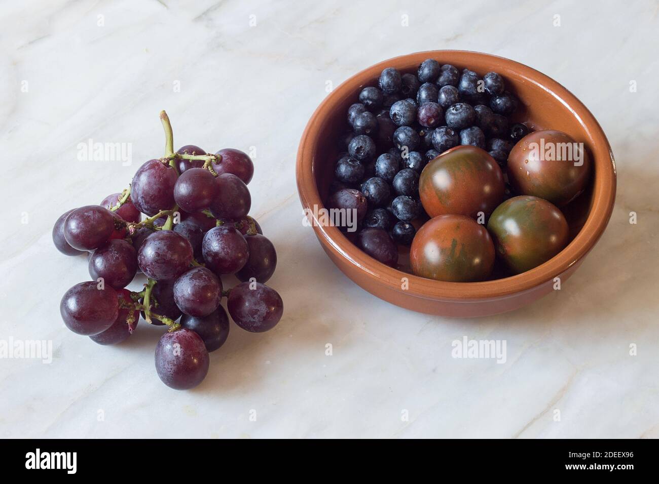 A rustic wooden bowl with blueberries and tomatoes from the garden next to a bunch of black grapes on an antique marble table. Healthy ingredients. Stock Photo