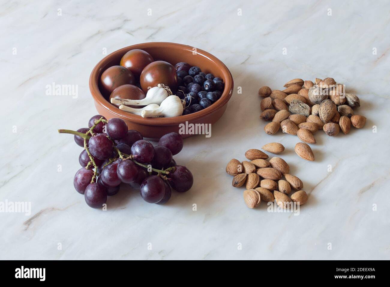 Several handfuls of natural almonds in the shell accompanied by a bunch of black grapes and a rustic clay bowl with blueberries, garlic, and tomatoes. Stock Photo