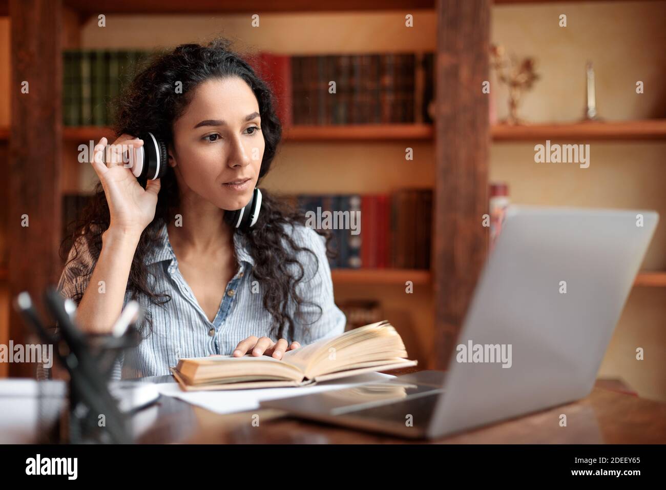 Woman sitting at desk, touching headset, using computer, reading book ...