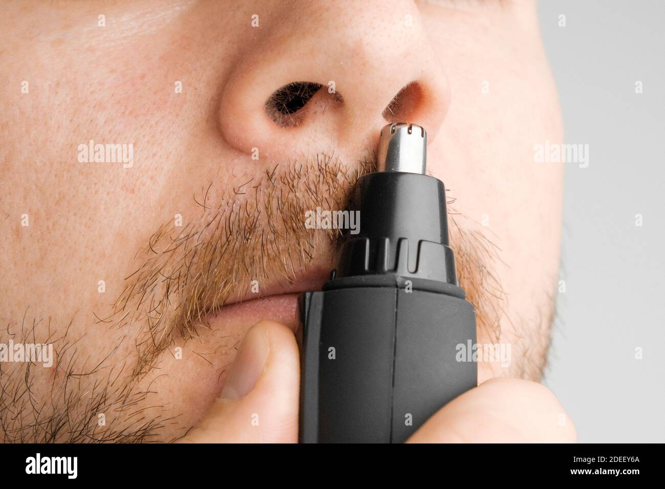Close up man removing hair from nose using electric trimmer on the grey background. Morning routine. Stock Photo