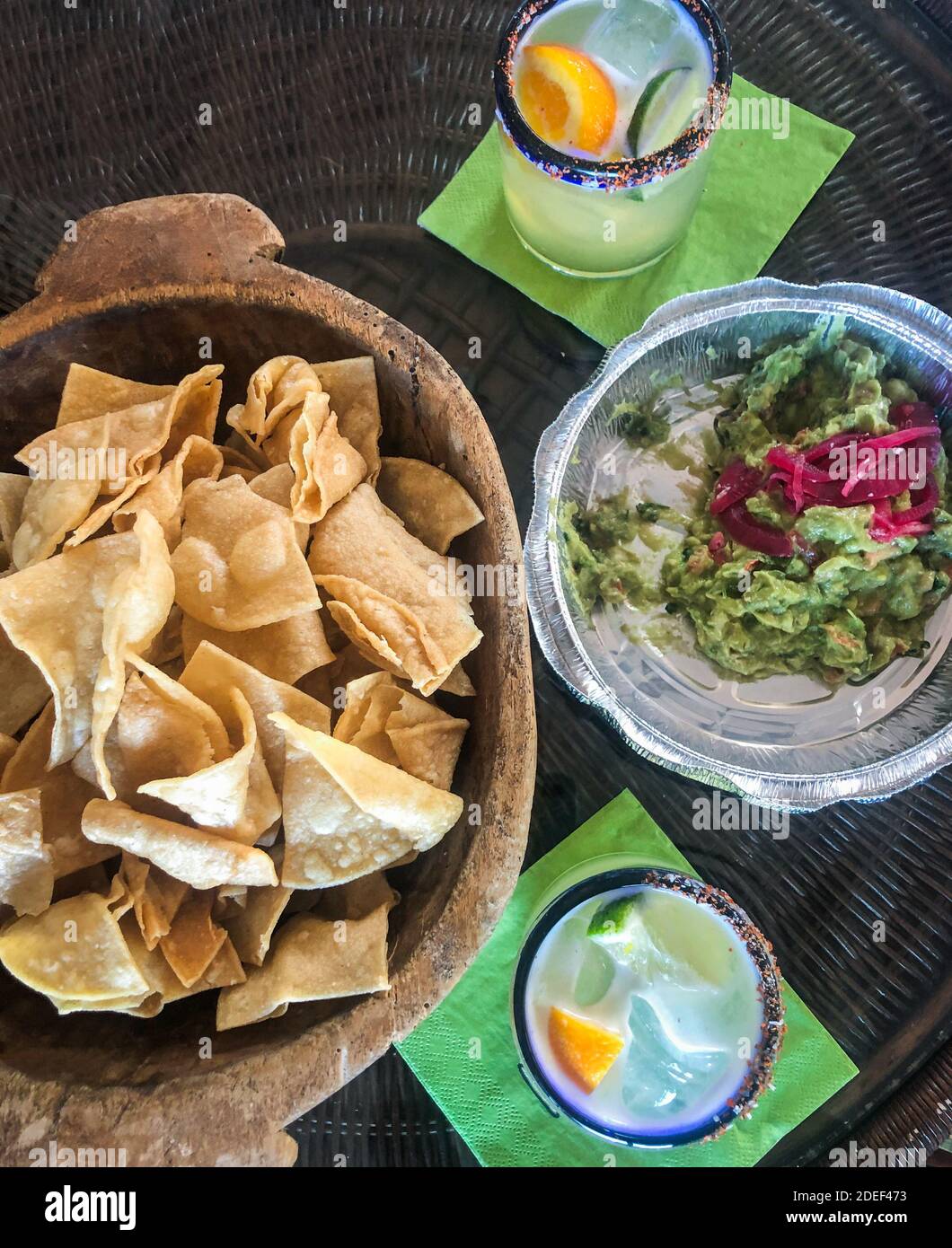 Mexican take-away food.  Take-out guacamole in a take-out container and chips in a wooden bowl with 2 margaritas in glasses with salted rims. COVID. Stock Photo