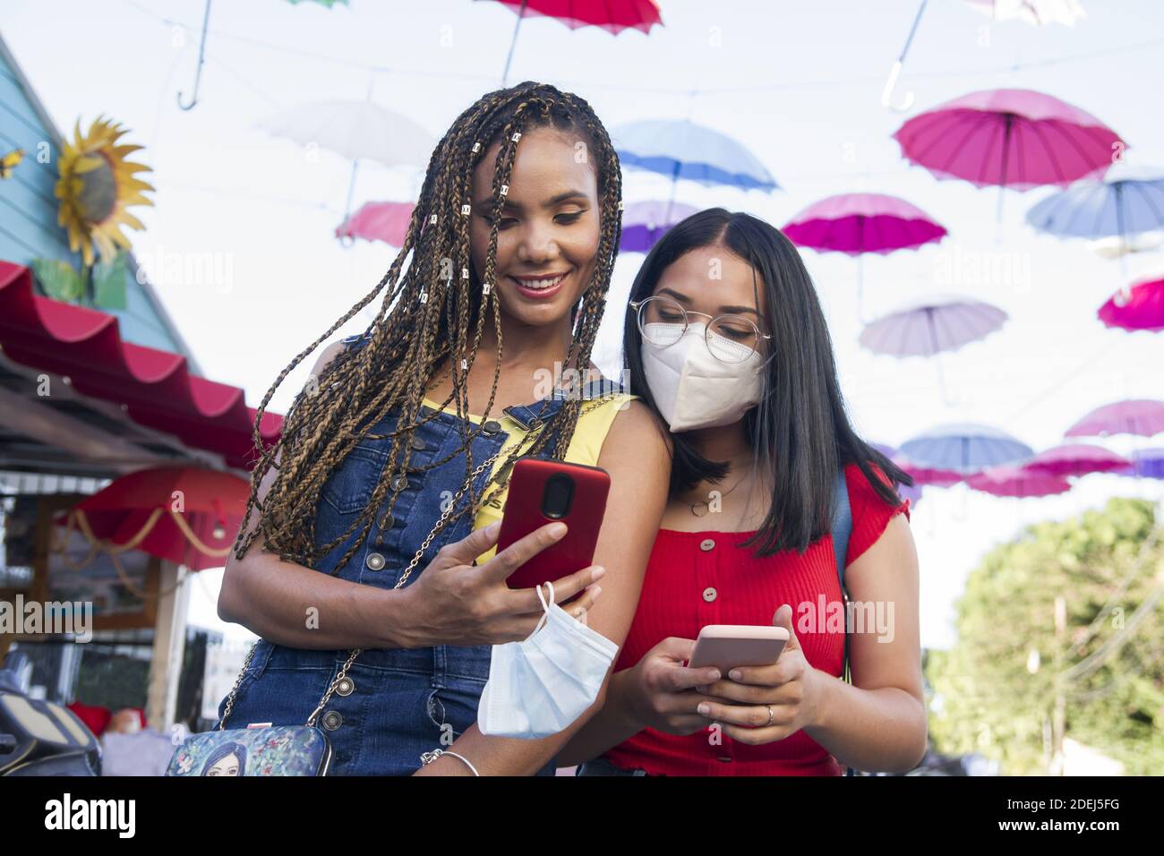 half body shot of two attractive young hispanic girls with face mask using smart phone in public street place in dominican republic with colorful umbr Stock Photo