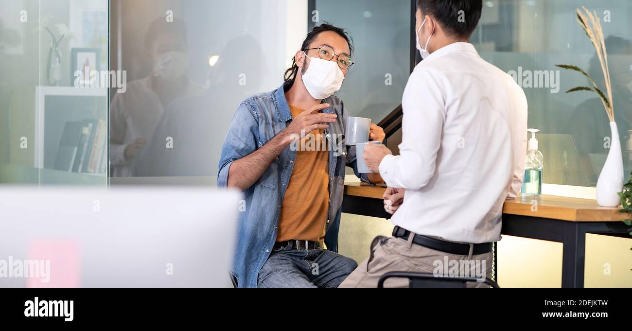 Panoramic Two office workers talking while coffee break in new normal with social distance practice office.  They wear face mask reduce risk to infect Stock Photo