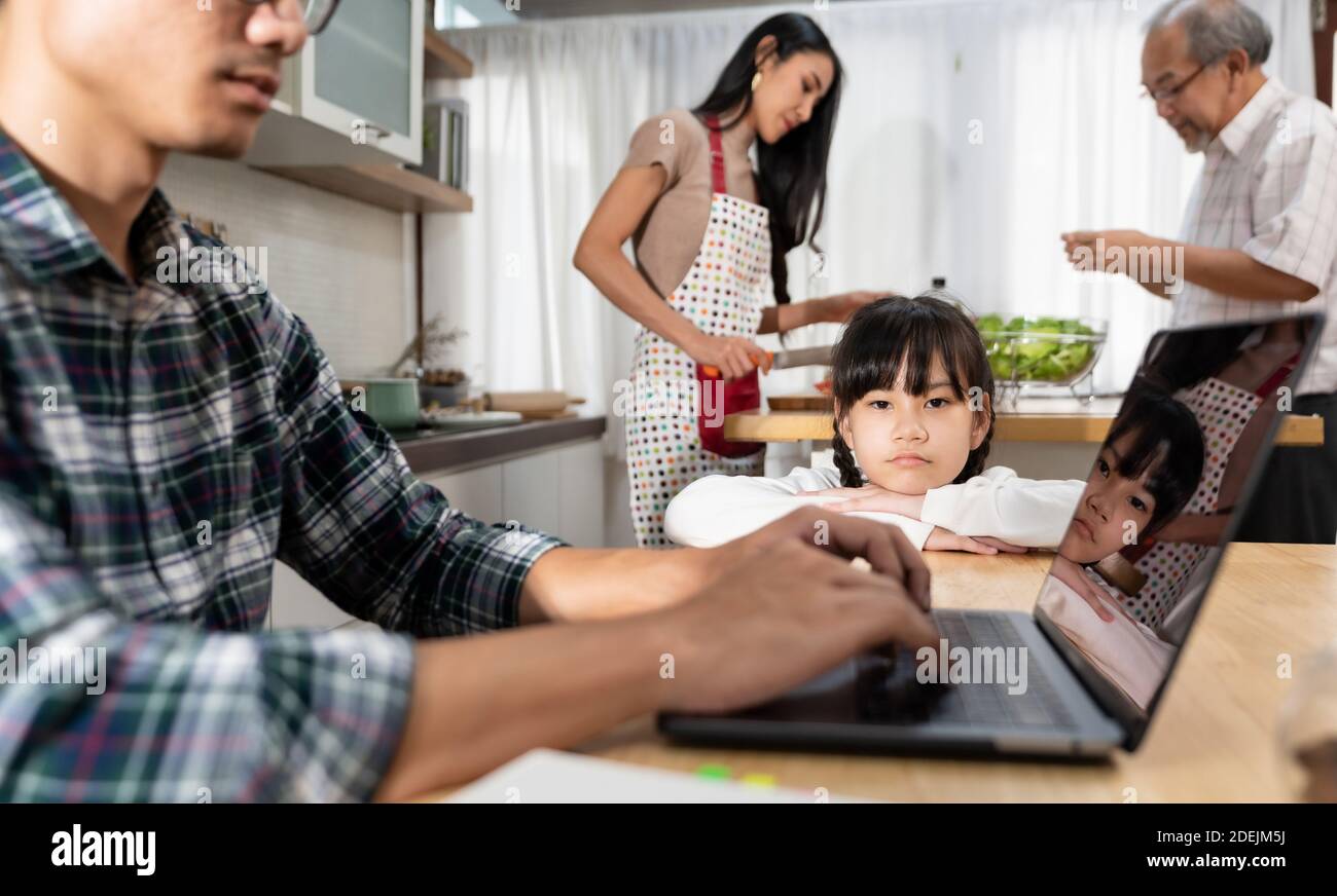Portrait Asian upset girl waiting her father working from home while city lockdown in dining room while mom and grandfather cooking in kitchen. Domest Stock Photo