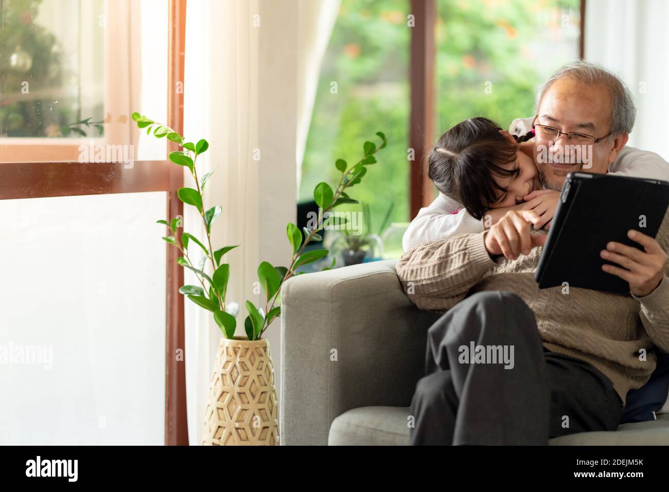 Happy retirement elderly man sitting on sofa at living room with granddaughter using digital tablet together. Multigenerational family with technology Stock Photo
