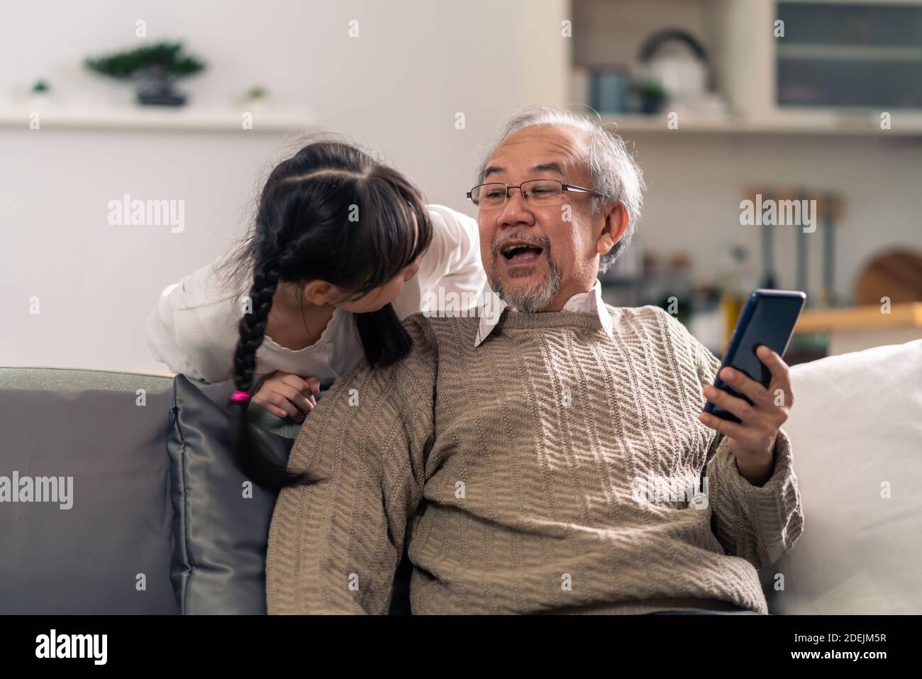 Happy retirement elderly man sitting on sofa at living room with granddaughter using digital tablet together. Multigenerational family with technology Stock Photo