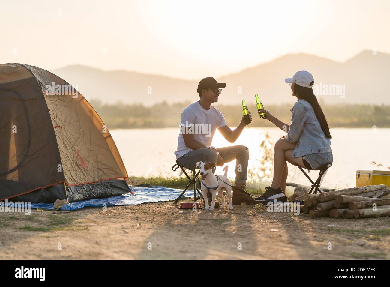 Young adult Asian couple drinking beer beside their tent campsite while sunset. Camping trip with dog outdoor activity campsite concept. Stock Photo