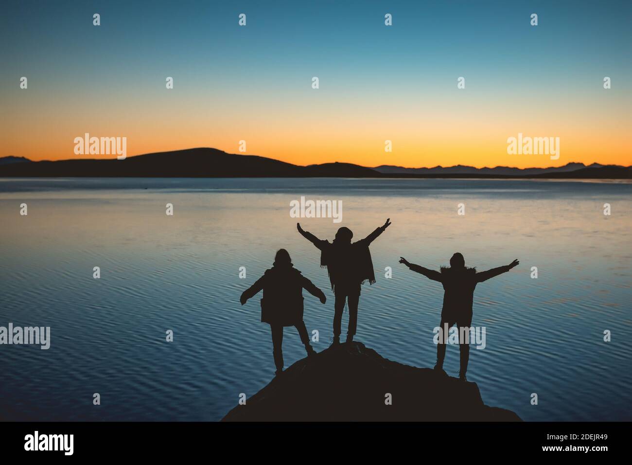 Silhouettes of three happy girls with raised arms stands against sunset lake and mountains Stock Photo