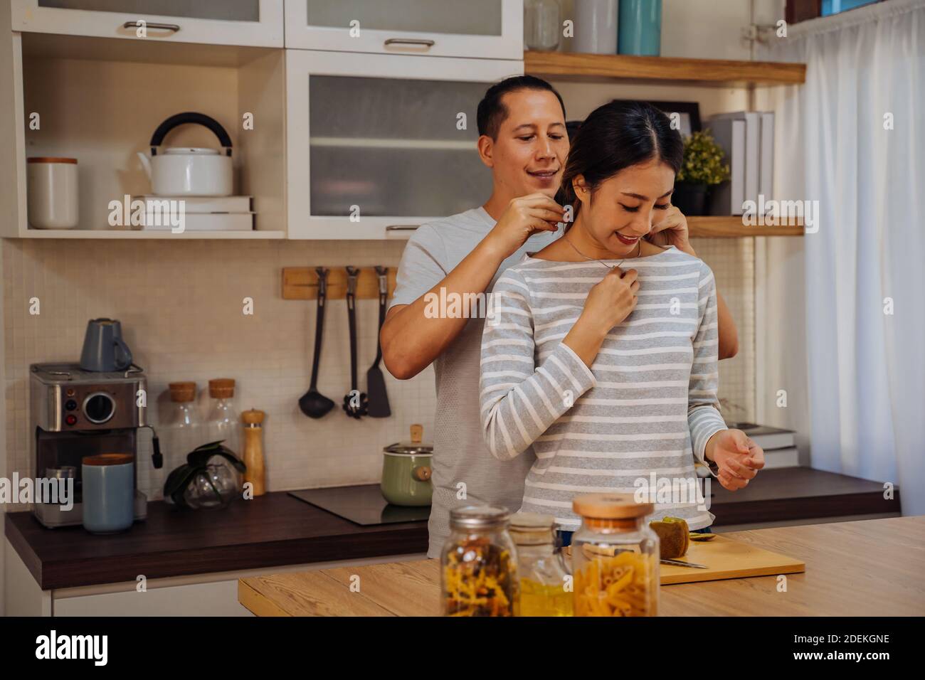Young man giving his girlfriend a necklace as present. Asian woman getting surprised by her boyfriend with marriage proposal while cooking in kitchen at home Stock Photo