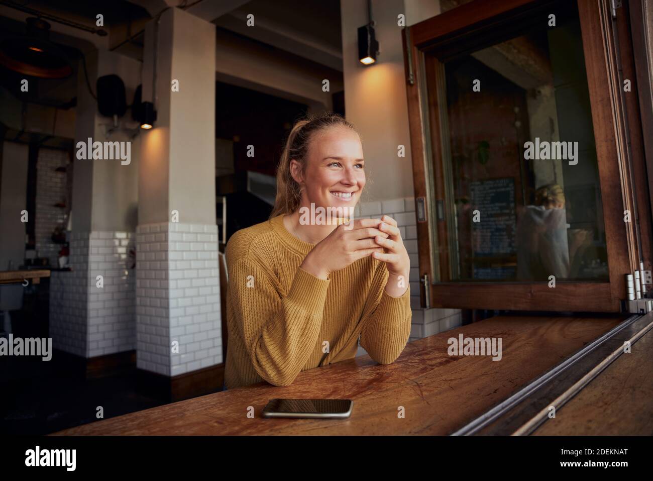 Cheerful female drinking coffee in cafe looking away with phone on table Stock Photo
