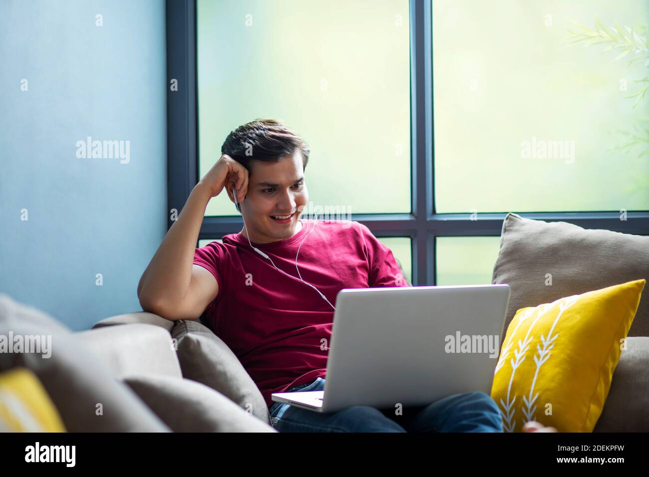 YOUNG MAN WATCHING A MOVIE ON HIS LAPTOP WHILE SITTING ON HIS COUCH COMFORTABLY Stock Photo