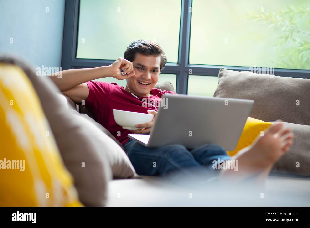 YOUNG MAN EATING CHIPS FROM A BOWL IN HIS HAND WHILE WATCHING A MOVIE ON HIS LAPTOP Stock Photo