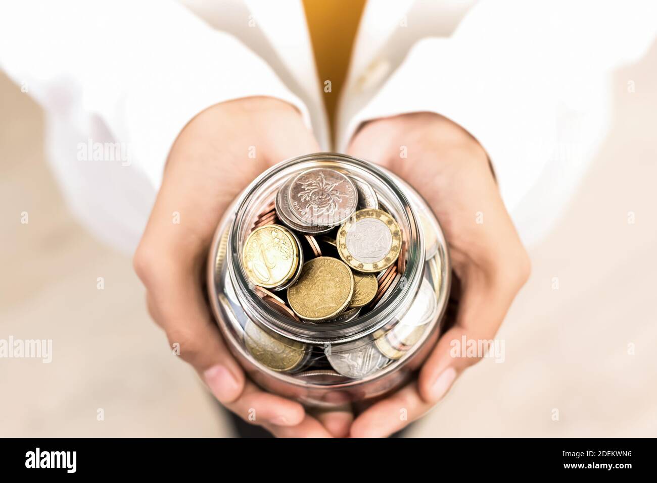 Young woman hands holding glass jar with multi currency coins inside, top view – money saving and financial concept Stock Photo