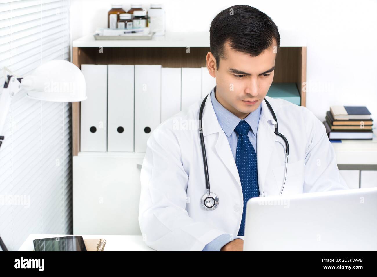 Handsome young Hispanic doctor working on laptop computer searching for information online in hospital room Stock Photo