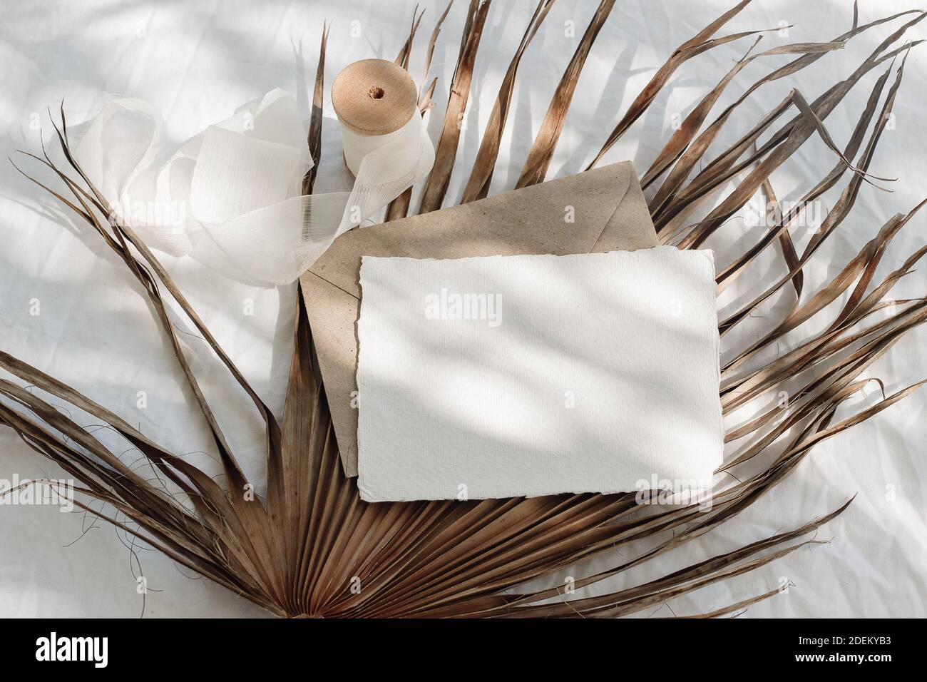 Tropical stationery still life. Closeup of blank card mock-up and craft envelope in sunlight. Dry palm leaf , white linen table cloth background Stock Photo