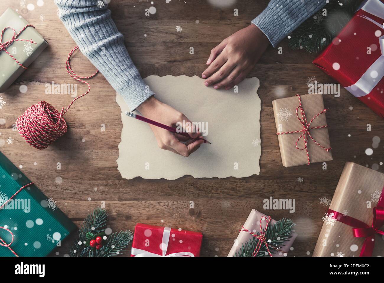 Woman in a gray sweater writing on craft paper with a red pencil in the center of christmas gifts on a wooden table with pine and mistletoe Stock Photo