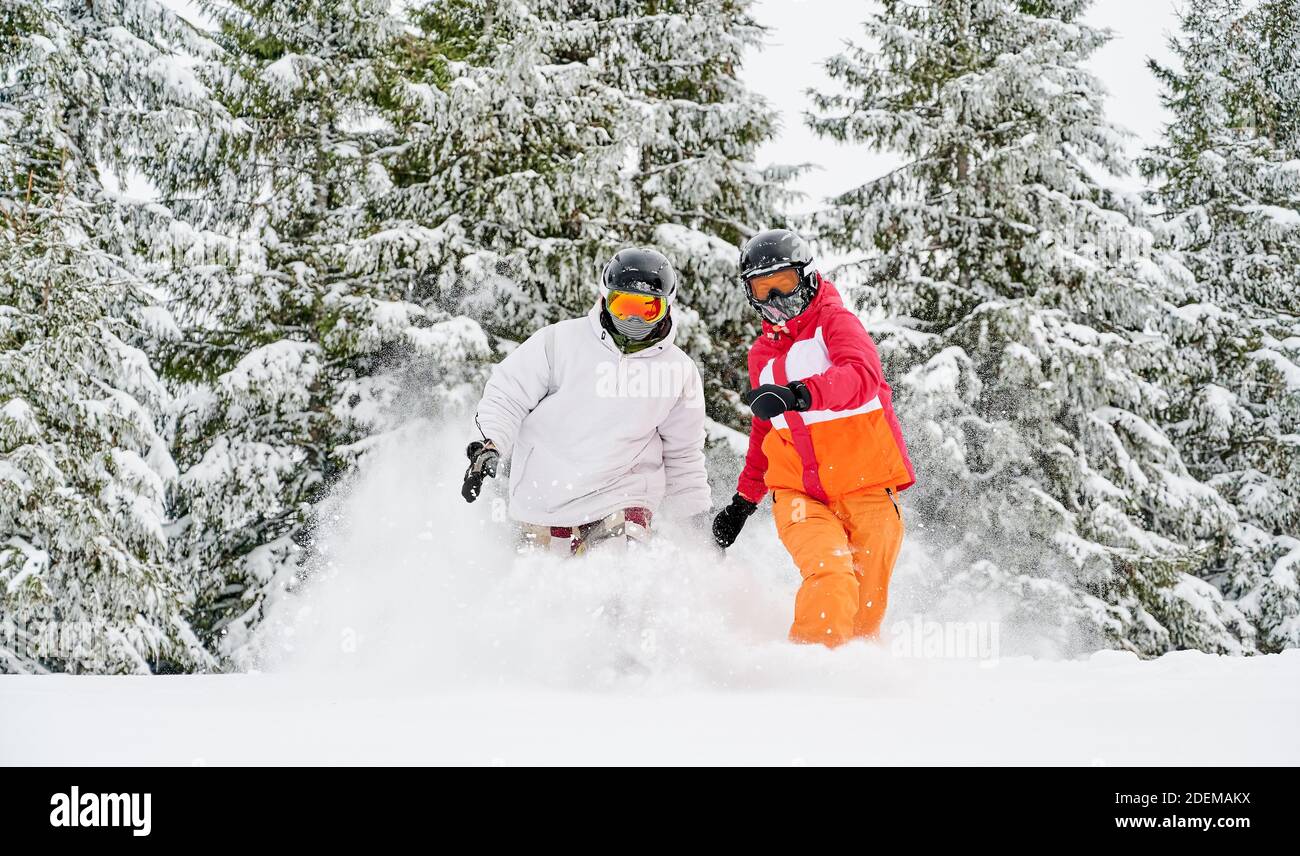 Man and woman in ski suits and helmets spending time together at ski resort, running through powder snow in winter forest with snowy coniferous trees on background. Concept of active leisure. Stock Photo