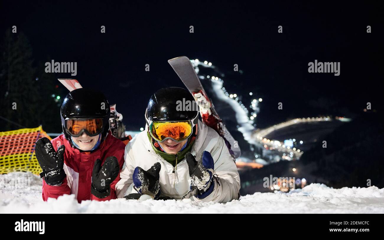 Couple of tourists lying on snowy slope at night, resting after active time spending on ski pistes. Close-up portrait of two cheerful happy people on snow Stock Photo