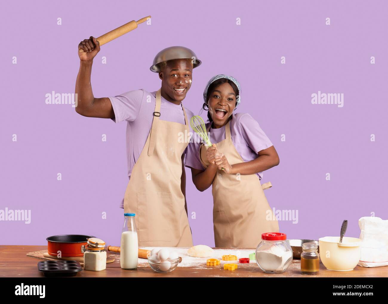 Funny black guy with girlfriend wearing aprons, holding kitchen utensils,  having fun while cooking on violet background Stock Photo - Alamy