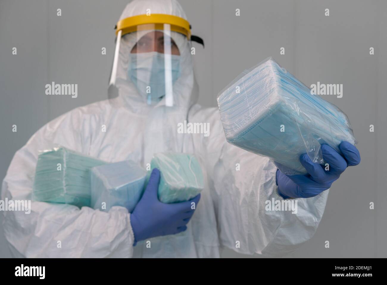 Male nurse wears protective suit with plastic face shield shows a pack of new medical masks during an pandemic of covid-19 in a hospital. Interior of Stock Photo