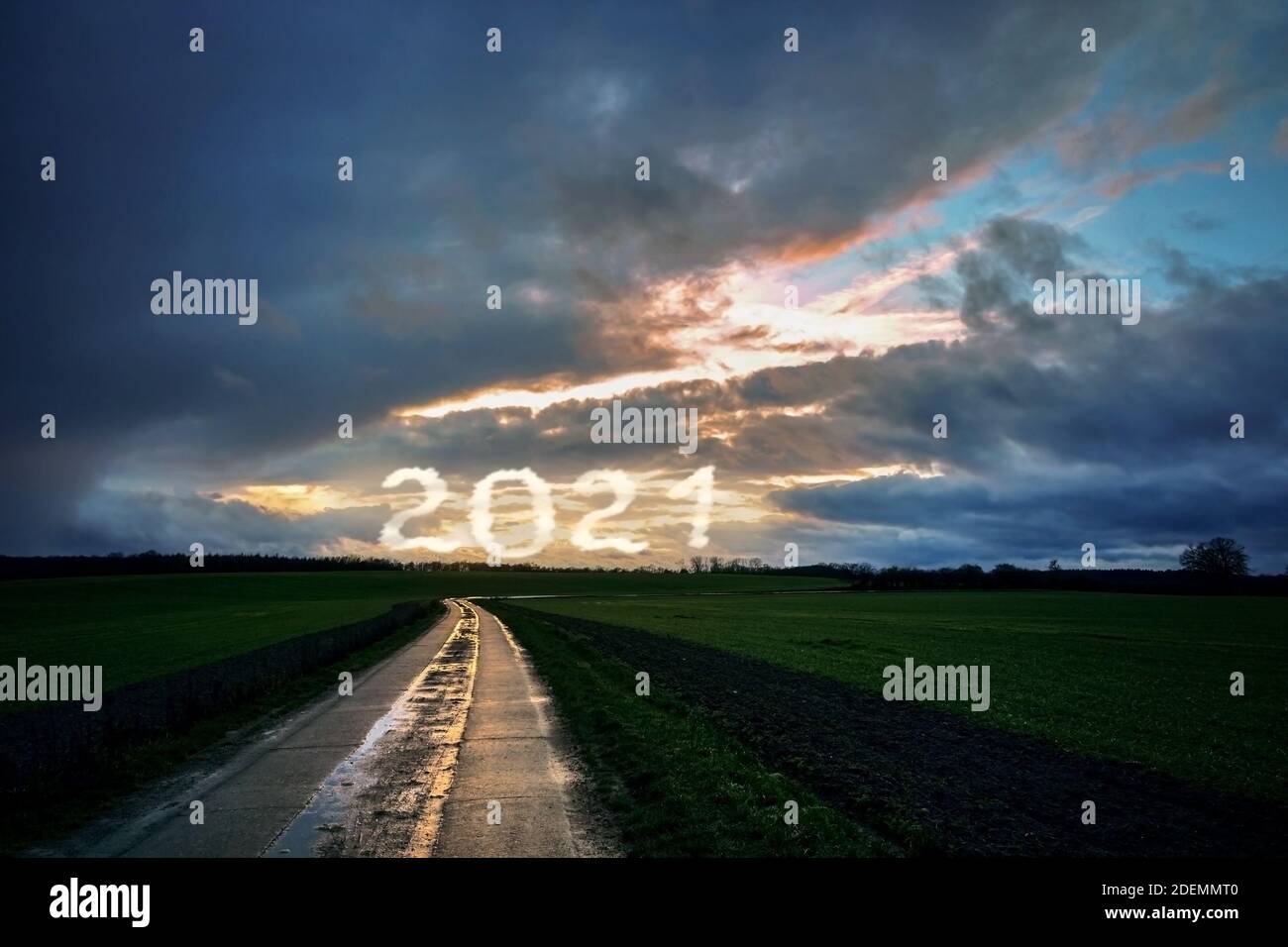 Wet reflecting country road is leading forwards through dark fields to a dramatic cloudy sky with New Year date 2021, future concept, copy space Stock Photo