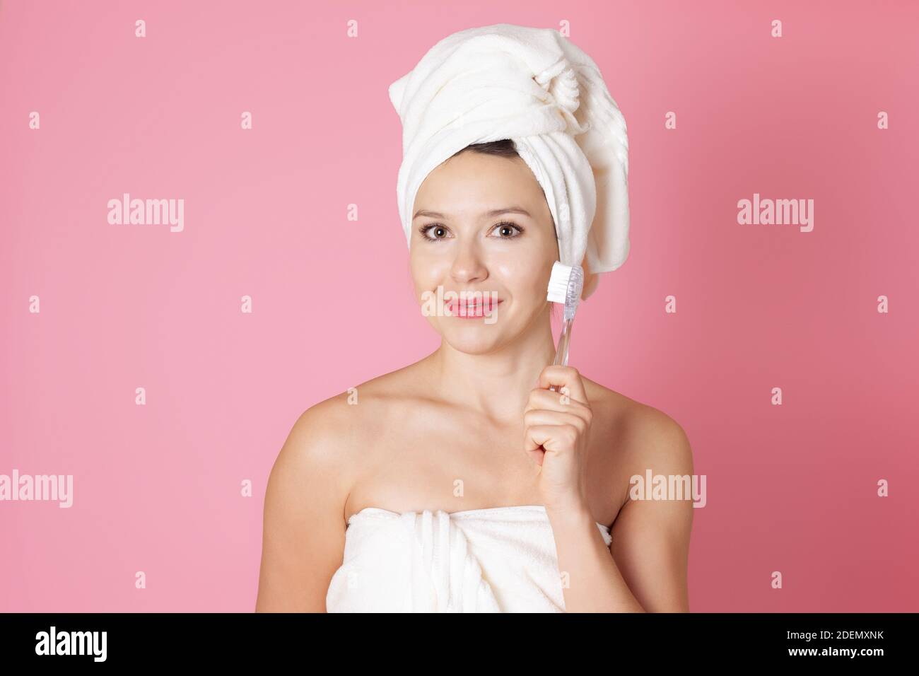 close up portrait of a young woman in a towel cleans her face with a glass brush and does an anti aging massage isolated on a pink background Stock Photo