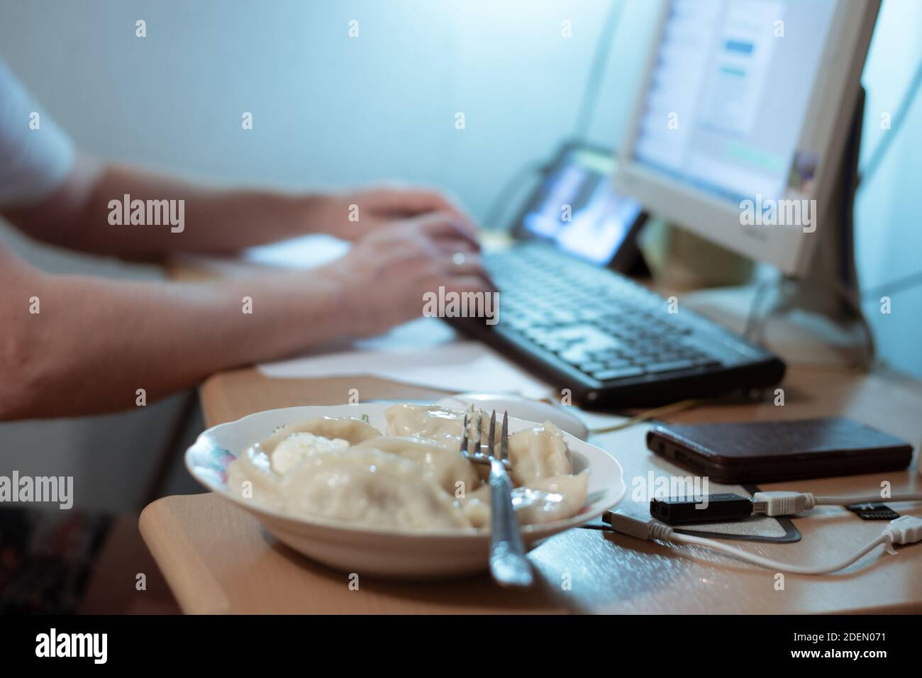 a man types on the keyboard at a computer table on which stands a plate of food, remote work at home during the epidemic Stock Photo