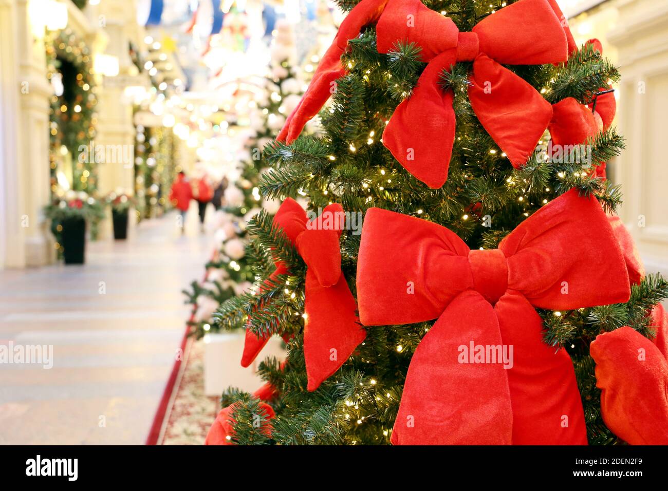 Christmas tree with red festive bows in a shopping mall on festive lights and walking people background. New Year decorations, winter holidays Stock Photo