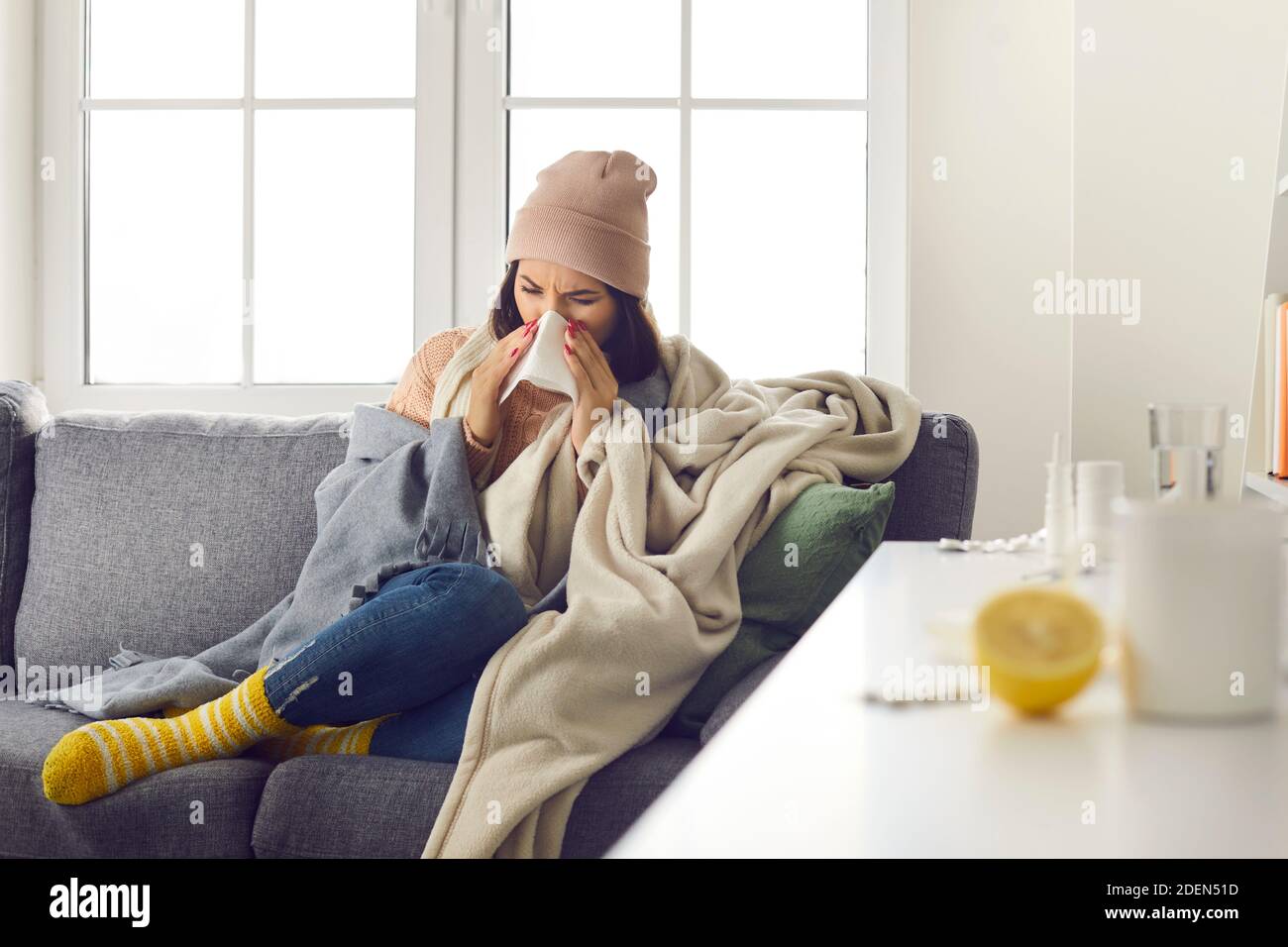 Young woman in warm clothing, socks and hat sitting on sofa, blowing nose and feeling ill Stock Photo