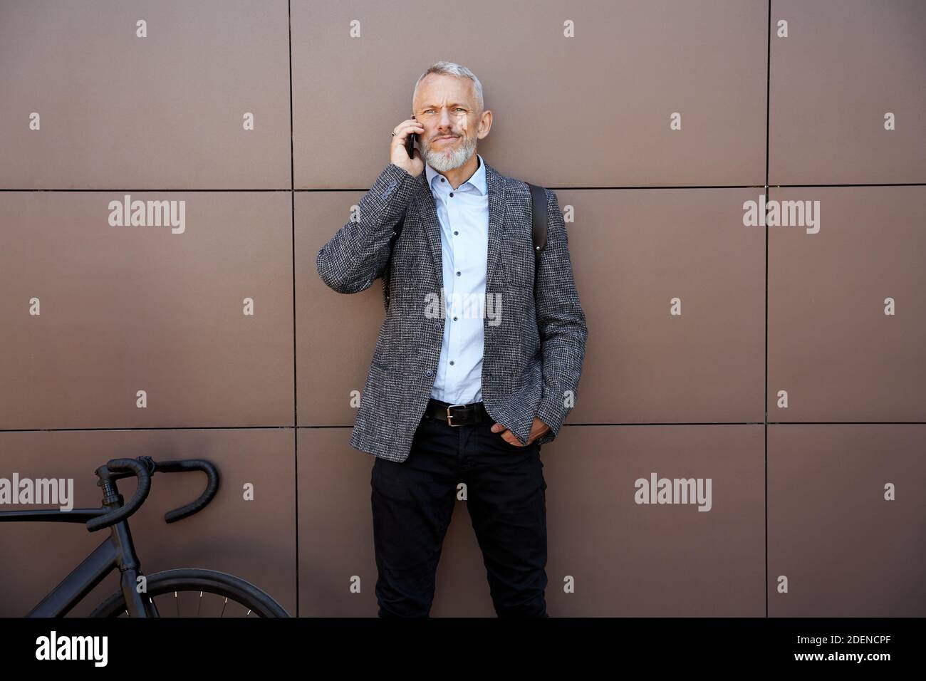 Modern middle aged businessman looking thoughtful while talking on the phone standing with his bicycle outdoors Stock Photo