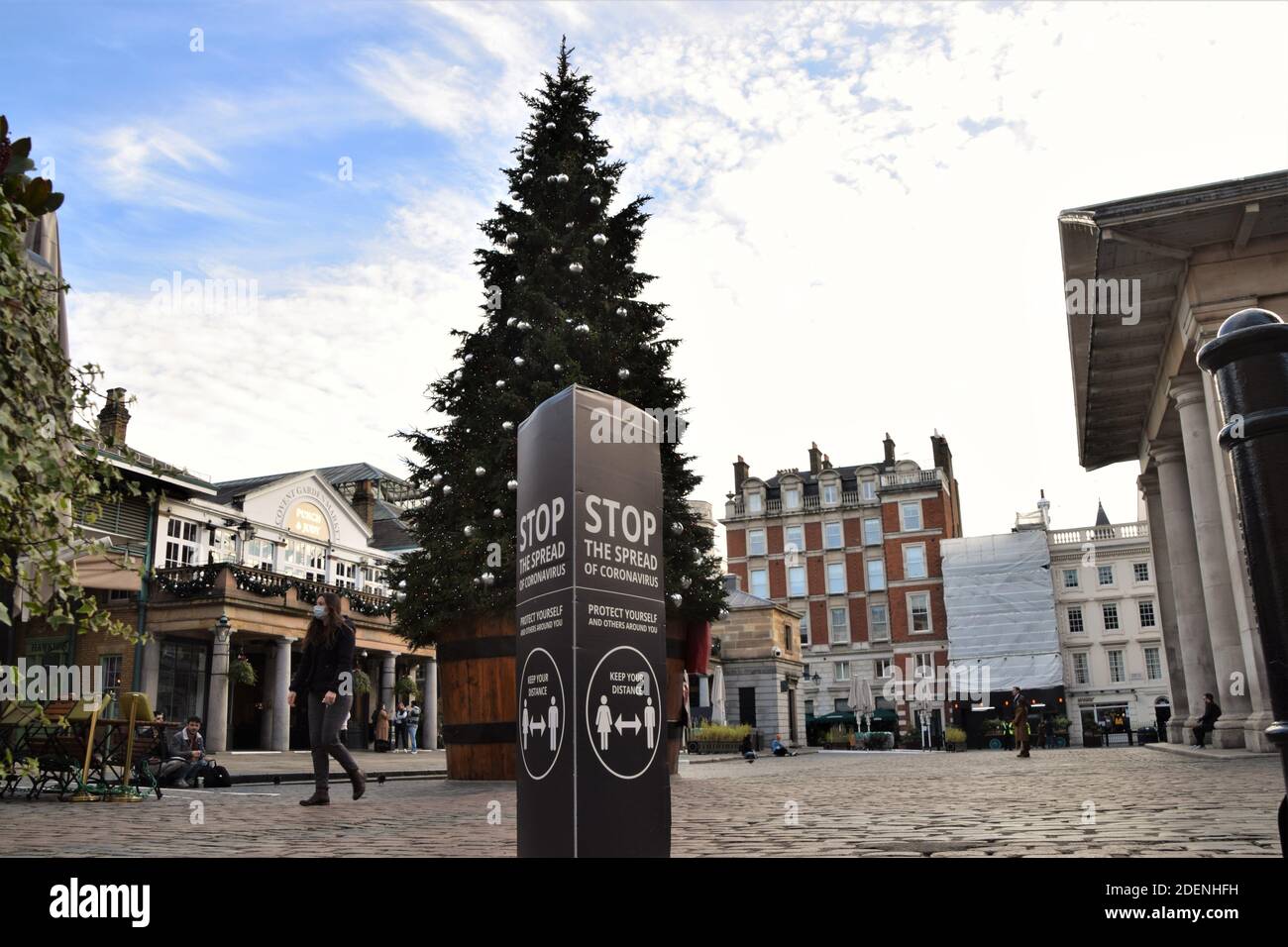 A woman wearing a protective face mask walks past a Stop The Spread Of Coronavirus sign and the Christmas tree in Covent Garden, London. Stock Photo